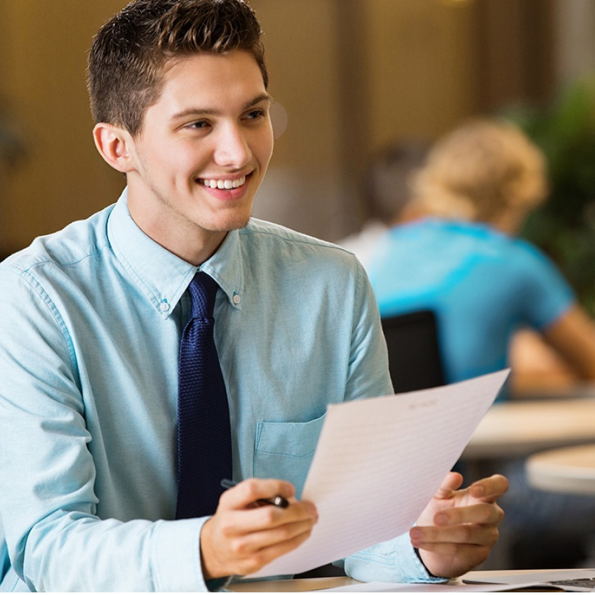Ein junger Mann mit Hemd und Krawatte hält ein Blatt Papier in der Hand und lächelt